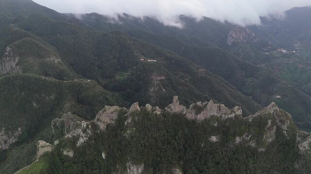 Aerial drone shot of the rugged, lush mountain landscape on Tenerife. The camera pushes in towards Roque la Monja, showcasing dramatic cliffs and dense forest under a cloudy sky.