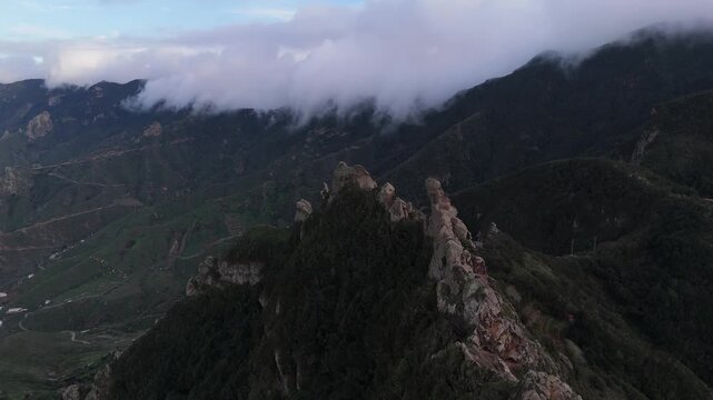 Aerial push in shot of Roque del Fraile mountain on Tenerife, Canary Islands. Lush, rugged landscape with dramatic peaks, dense forest, and distant village under cloudy skies.