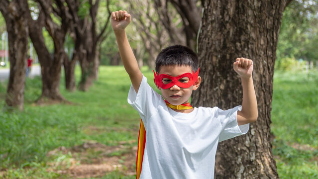 A happy little Asian boy wearing a red superhero mask raising both fists with excitement and enthusiasm while playing pretend games in a sunlit public garden. High quality stock photo