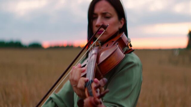 Woman playing violin in golden field at sunset, focusing on graceful hand movements and emotional connection with music in a serene rural landscape