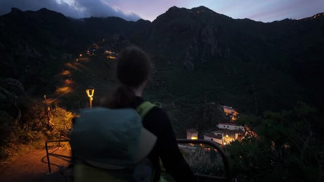 A person hikes a winding trail down to Afur on Tenerife, Canary Islands, as twilight descends over rugged mountains. Distant village lights dot the lush landscape, offering a serene evening view.