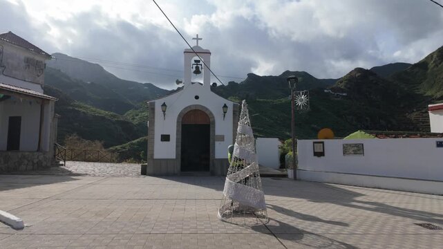 A lone hiker explores a picturesque, remote village of Afur nestled amidst rugged green mountains on Tenerife. The charming white church stands out under a sunny, cloudy sky, casting long shadows.