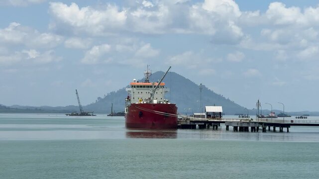 Large industrial ship loading cargo at a terminal with hills in the background under clear skies