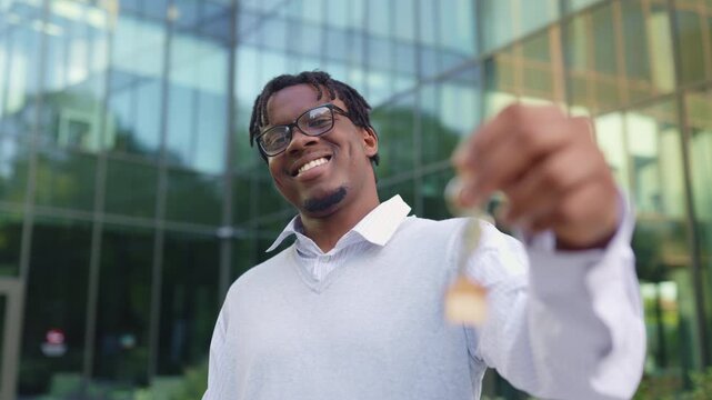 A smiling african american man proudly holds up a set of keys with a house-shaped keychain in front of a modern glass building
