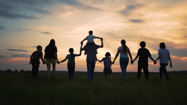 Large family silhouette walking through open field at sunset. Group is holding hand walking together. Happy family enjoys beautiful nature. Small child rides parent shoulder. Sunset lights group.