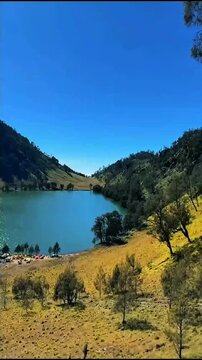 Bright clear blue sky hangs over calm turquoise lake, grassy golden hills and forested slopes surround shore with small camping settlement