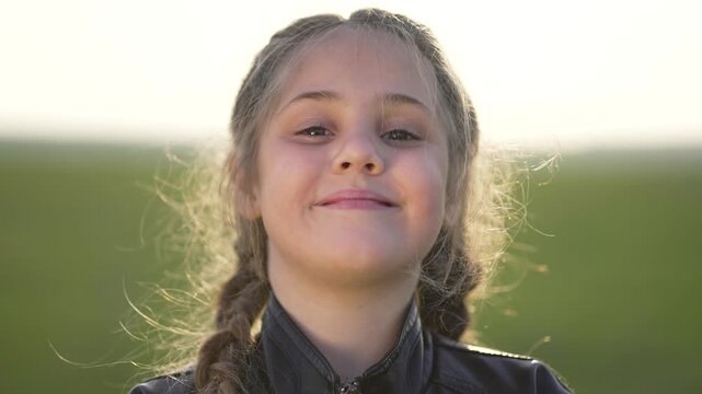 Little girl face close-up in green field at sunset. Child with braid looks at camera. Girl portrait shows emotion from serious to smile. Kid face glows in warm light. Happy child smiles in field.