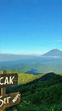 Sharp volcanic cone rises above rolling green forested hills, clear bright blue sky stretches wide, soft mist veils distant valleys below