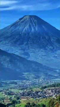 Massive steep volcanic mountain towers over clustered village homes and green farmlands under a vivid clear blue sky with wispy clouds