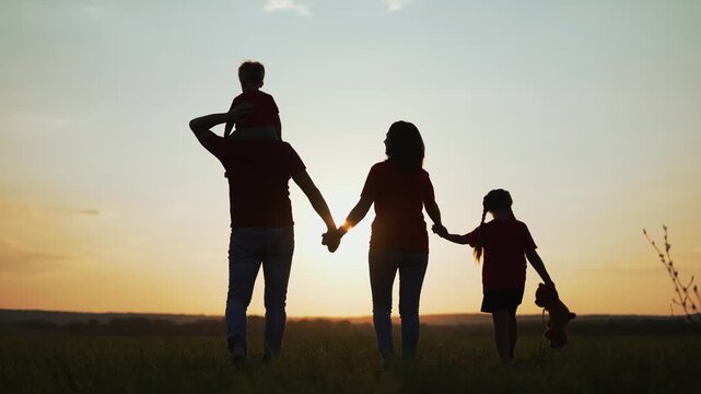 Family walks together at sunset in field. Father carries son on shoulders. Mother holds daughter hand. Daughter holds teddy bear. Family silhouette glows against sun. Happy family walk outdoors.