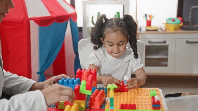 Girl placing a plastic block with hands on a colorful table in a studio playroom with tent and toy kitchen in view; concentration learning play.