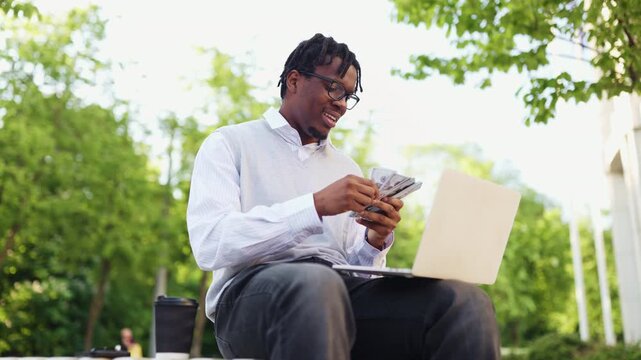 A young african american man wearing glasses and a vest sits outdoors with a laptop and counts a stack of US dollar bills