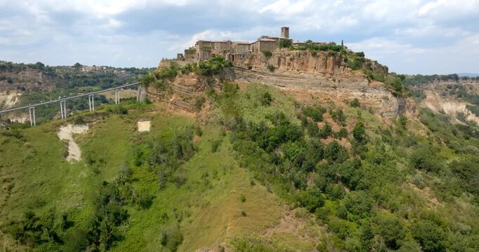 Panorama of Civita di Bagnoregio, Italy,  showing stone buildings atop steep tufa cliffs, connected by a long pedestrian bridge, under a partly cloudy sky with green hills in the surrounding landscape