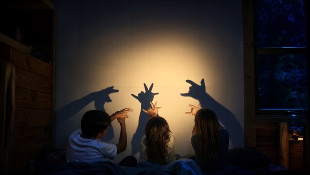 Three children making shadow puppet on wall in dark wooden cabin bedroom. Child playing with light and shadow at night during summer holiday sleepover. Imagination and fun