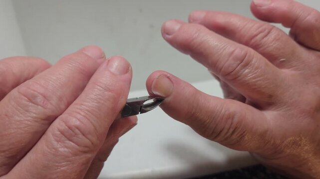 POV shot of an elderly woman using sharp metal cuticle nippers to precisely trim hard overgrown skin and excess cuticle growth along the side edge of her index finger during a detailed manicure.