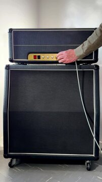 Vertical video social media - A music performance valve amplifier and speaker cabinet, standing indoors, with a man&rsquo;s hand plugging in an instrument cord, lead or cable, then switching the power on.