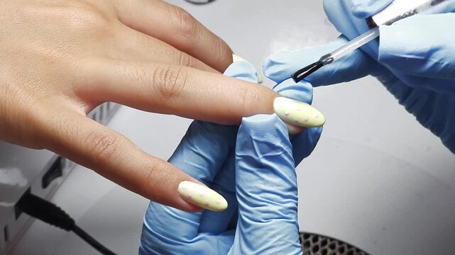 Russian manicure finishing with top coat. Professional nail technician applying glossy top gel over pastel polka dot nail design. Close-up beauty salon procedure.