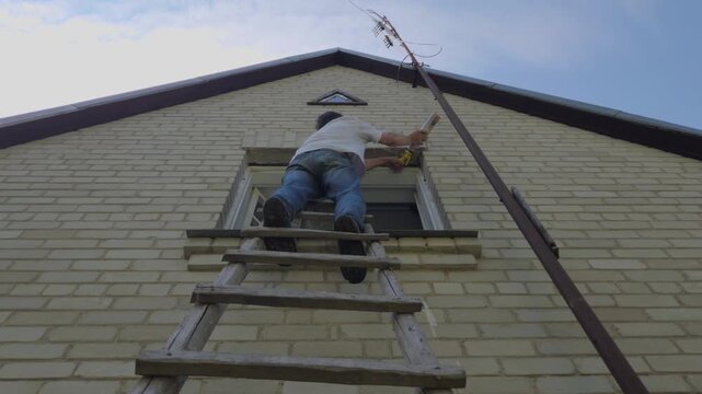 Low-angle shot of a man standing on a wooden ladder using polyurethane expanding spray foam to seal and insulate the gap around a second-story window frame on a brick house wall.