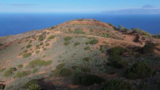 Static aerial drone shot capturing a rugged, remote island landscape on La Gomera (Canary Islands), featuring hills, lush vegetation, and the ocean with the Teide of Tenerife under a sunny sky.