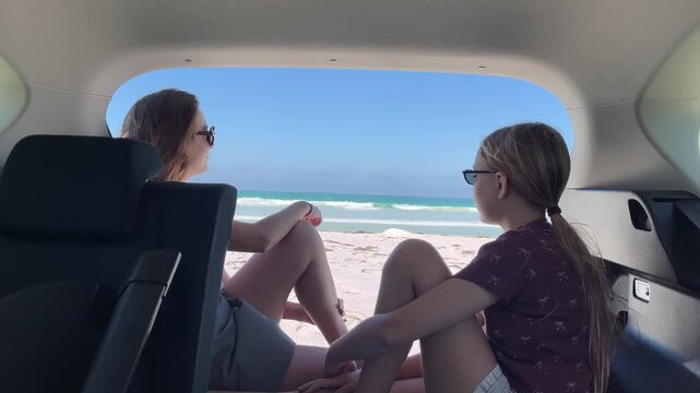 Mother and daughter sitting together in the trunk of a car overlooking the beautiful sandy beach and ocean during the daytime.