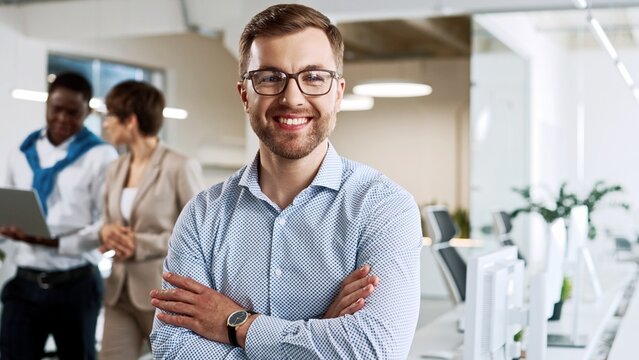 Cheerful young Caucasian man in glasses standing in cabinet in modern company looking at camera and smiling. Close up of joyful handsome male manager at workplace. Office worker. Business concept