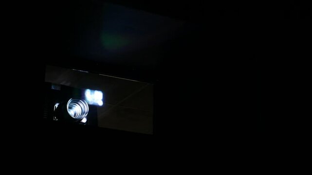 Bright beam shine through projector lens and protection glass, dark movie theater room. Small particles fly in air, highlight by imager light. Blurred picture reflex on window glass in front of lens.
