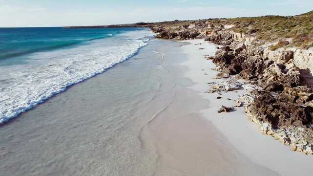 Ocean waves wash over the white sand beach along the rocky cliffs during a bright day on the coast of Western Australia.