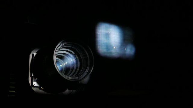 Cinema projector lens and blurred movie image on glass, closeup view. Some motion and illumination change, modern projecting camera optical front end, large diameter lens. Image reflecting on operator