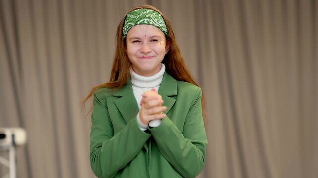 Teenage girl in green blazer scrunches up her face and rubs her hands together, acting like a comic villain during school drama rehearsal.