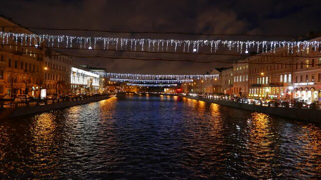 Fontanka river perspective at night, unfrozen water, new year garland on tensed rope across, old buildings, many lantern lights and lams glow. Reflection and small waves ripples over water surface,
