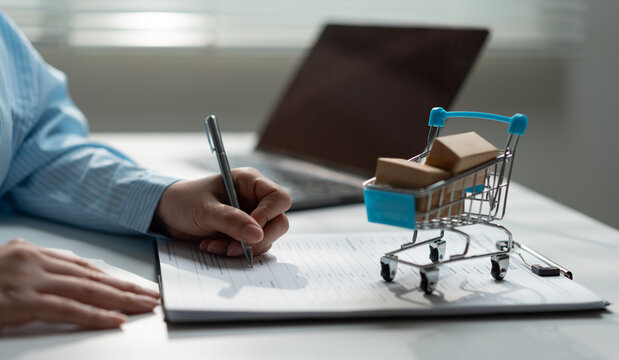 Business professional signing documents beside shopping cart with parcel boxes, representing purchase verification, ecommerce documentation, quality approval, procurement and retail management.