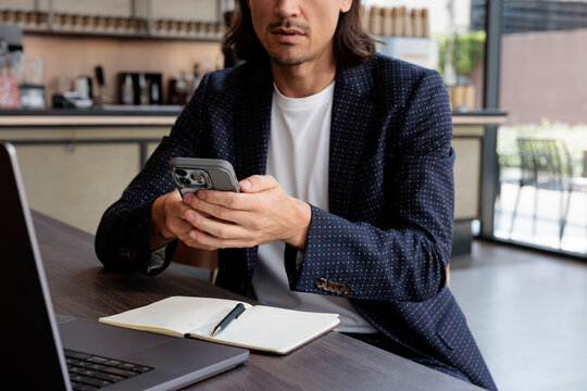 Man in blazer uses mobile phone at desk with laptop.