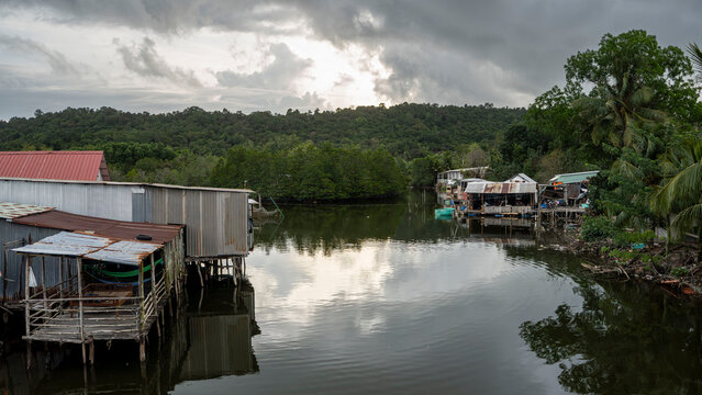 A quiet Vietnamese fishing village with weathered stilt houses lining a calm waterway, flanked by mangroves and dense jungle hills under a dramatic overcast sky.