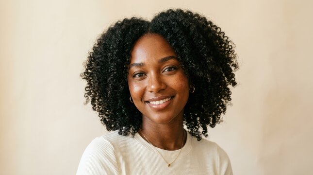 A woman with curly hair smiling, wearing a white shirt and a gold necklace, standing in front of a beige background.