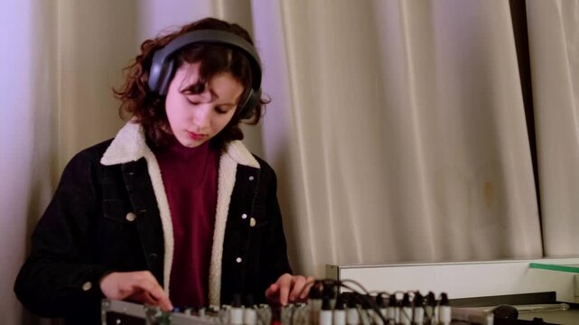 Young girl in casual jacket works carefully at a mixing console with headphones on, creating modern electronic music and developing creative technical skills.