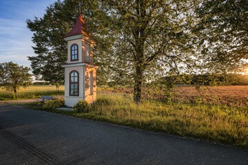 Naklejka premium A traditional wayside shrine chapel standing by a rural asphalt road and a blooming field during sunset in the countryside.