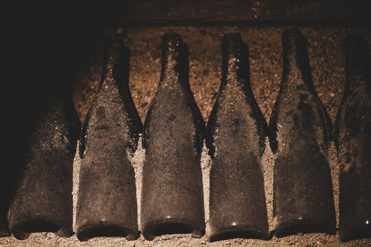 A row of aged wine bottles covered in dust rests on a sandy surface inside a dimly lit cellar, symbolizing long-term preservation.
