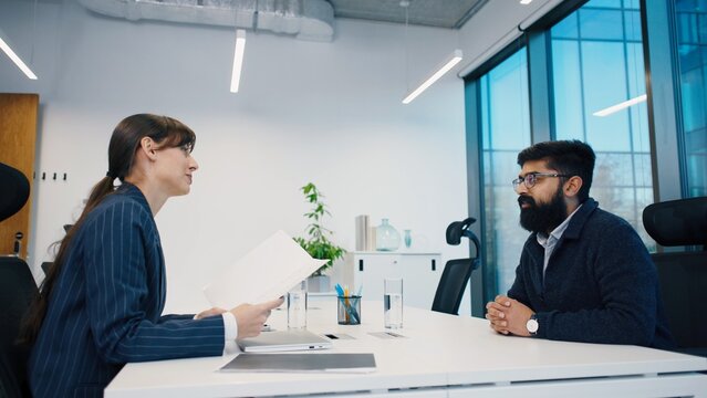 Caucasian HR woman discussing work during job interview. Man holding portfolio, smiling politely. Bright glass office setting, modern business environment. CV interviewing concept. Applying for work.