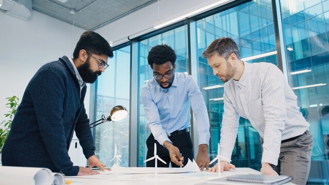 African American male explaining concept to South Asian and Caucasian colleagues during discussion in office. Three professional men planning wind energy model project near window. Indoors.