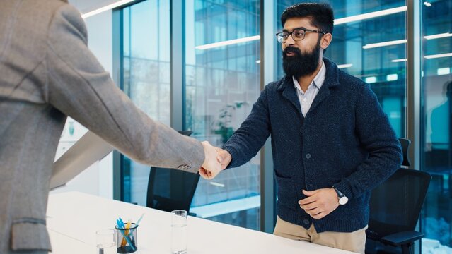 South Asian man with beard attentively answering questions during job interview with Caucasian man holding folder. Business setting with glass walls and professional tone. Males shaking hands.