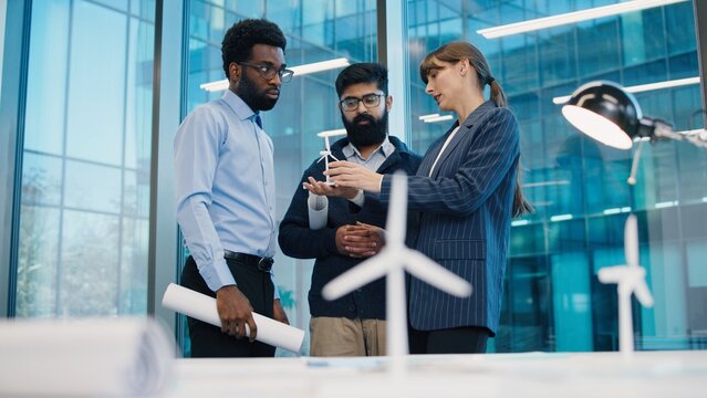 Caucasian woman showing wind turbine model to African American and South Asian colleagues in modern office. Professionals discussing sustainable energy prototype during meeting near window. Indoors.
