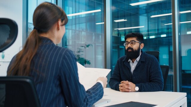 Indian man with beard and glasses sitting confidently during interview, Caucasian HR woman holding resume, discussing qualifications in modern glass office. Female boss interviewing new employee.