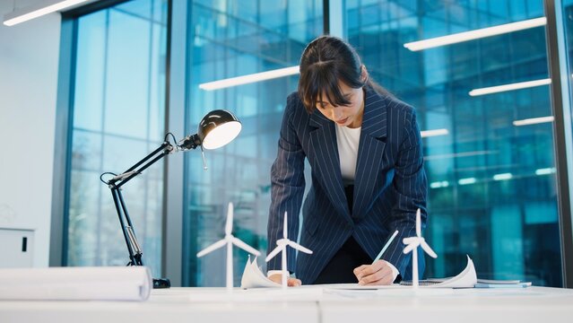 Caucasian woman working with blueprints in modern office. Female engineer or architect planning project near wind turbine models. Indoors. Business environment. Office work concept.