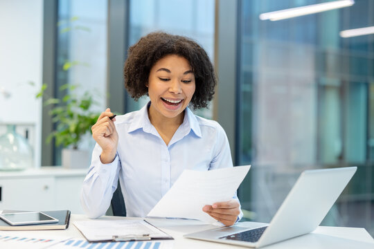 Happy young African American businesswoman sitting at the desk in the office, reading documents and rejoicing, showing a victory gesture with her hand