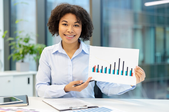 Portrait of a smiling young African American woman sitting at a desk in the office and showing documents with graphs to the camera.