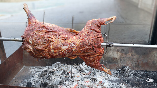 Whole lamb roasting on a metal spit above glowing charcoal during traditional outdoor barbecue. Authentic slow cooked meat prepared over an open fire with rich golden roasted texture.