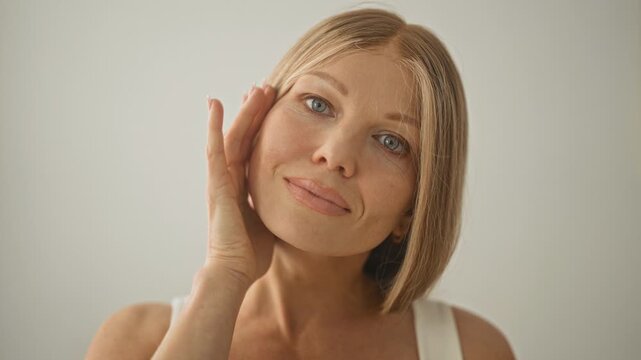 Woman gently touches cheek with fingertips, head tilted and eyes to camera in white studio with soft natural light; serenity wellness.