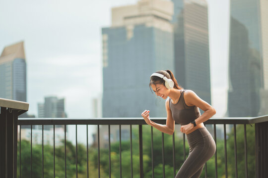 Excited Asian sportswoman celebrating success fist pump while running at city park bridge, cheerful fit female runner wearing headphones enjoying outdoor workout for urban health and active fitness
