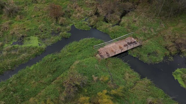 Dilapidated and disused, an old bridge remains resolute in its task of spanning a small creek. Gentle, smooth water flows beneath and between lush green in a boggy wetland. Aerial footage of a waterwa