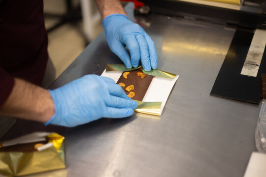 Hands in blue protective gloves carefully packaging a handmade chocolate bar with nuts in a small paper box. Close-up of artisanal chocolate production, confectionery preparation and hygienic food